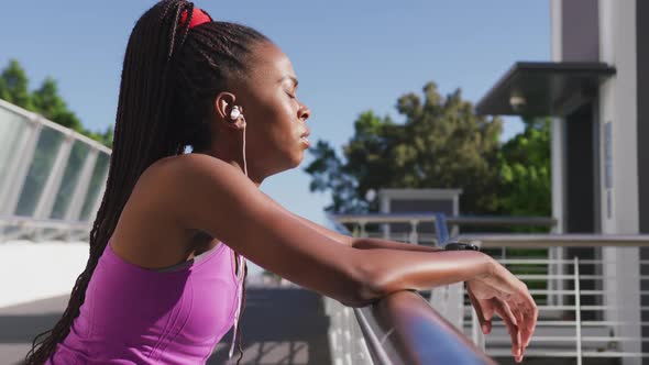 Tired african american woman wearing earphones taking a break from running on the city bridge