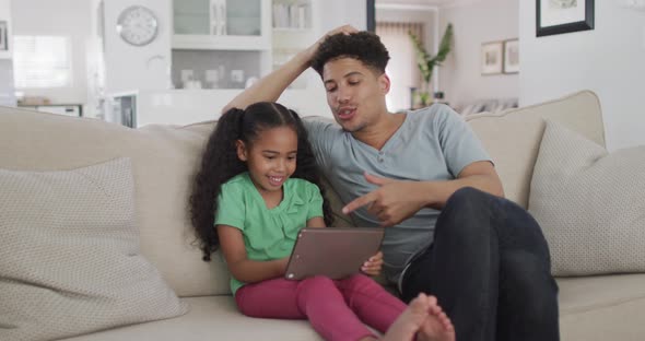 Happy biracial father and daughter sitting on sofa using tablet
