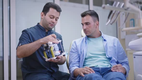 Concentrated Caucasian Man Listening Male Middle Eastern Dentist Talking Showing Dental Cast
