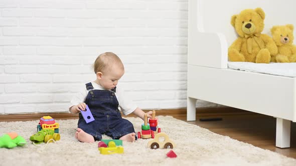 Baby Boy in Denim Overalls Plays with Toys Sitting on the Carpet