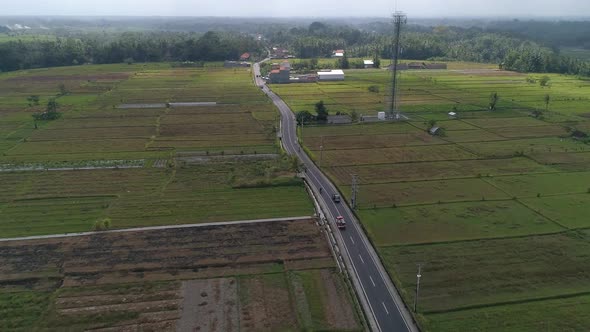 Aerial view, cars travelling down Bali road surrounded by farmlands. High angle pan right.