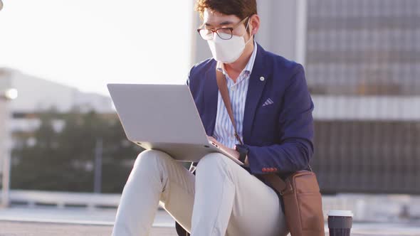 Asian man wearing face mask using laptop while sitting on the stairs at corporate park