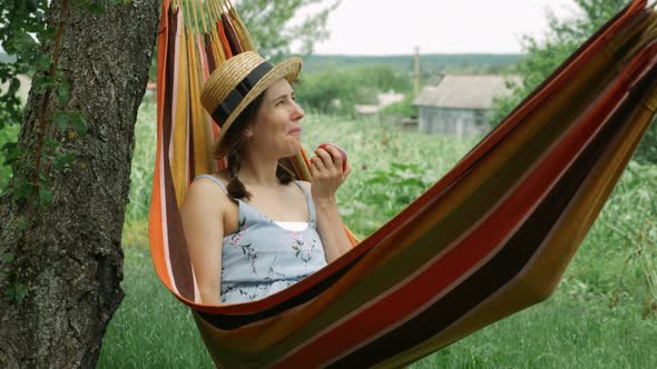 Young woman lying in hammock and enjoying summer vacation at countryside eating apple
