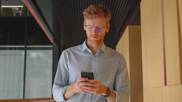 Young Adult Smiling Businessman Texts Messages on Mobile Phone App in Office