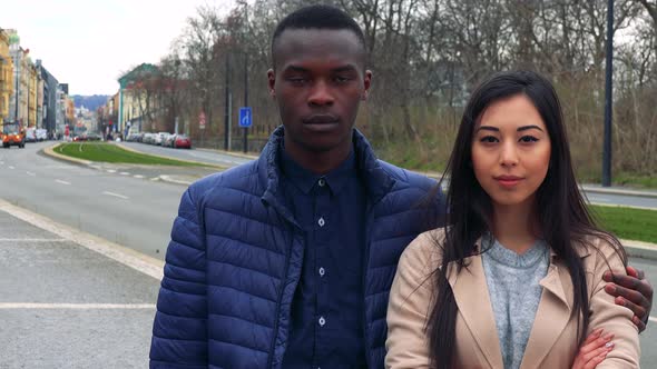 A Black Man and a Young Asian Woman Look at the Camera on a Sidewalk, His Arm Around Her Shoulders