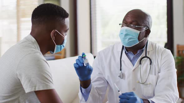 African american senior male doctor giving covid vaccine to male patient in home, wearing face masks