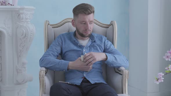 Happy Thoughtful Rich Bearded Man in Blue Shirt Sitting in the White Armchair in the Light Room