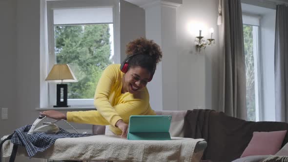 Positive Young African American Woman Ironing Laundry with One Hand and Talking at Tablet Webcam