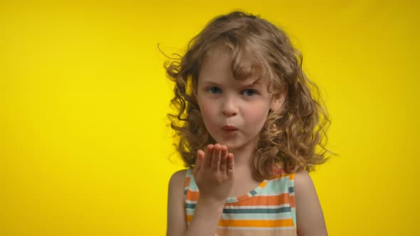 Beautiful Little Curly Blond Girl in a Striped Dress is Blowing a Kiss