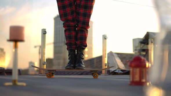 Legs of Unrecognizable Slim Woman in Checkered Pants Standing on Skateboard with Urban Skyline at