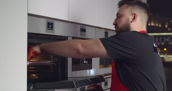 Young Man in Apron Taking Tray with Meal From Electric Oven in Kitchen