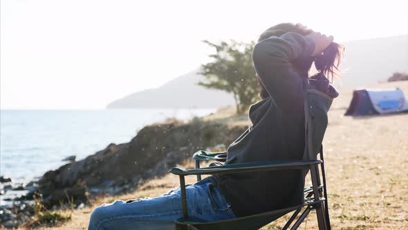 Young Man Sits on Chair at Campsite on Lake Shore and Collect His Hair in Bun