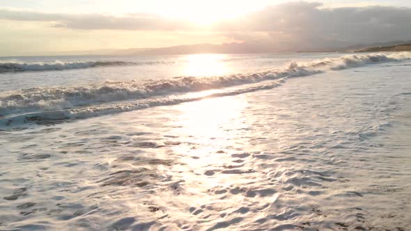 Aerial upward flight over waves crashing at beach at sunset while getting close to water level SLOW