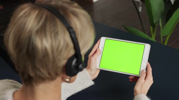 A Middleaged Woman with a Headset is Viewing Content Using a Tablet on a Green Screen Calling Her