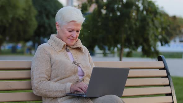 Aged Woman Sitting on Bench in City Park Using Computer Surfing Internet Smiling Senior Female of