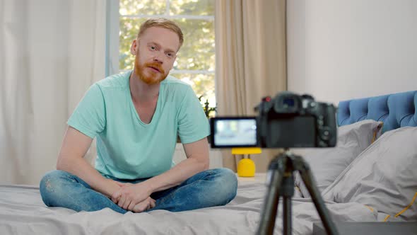 Young Man Blogger Recording Video Blog on Camera and Talking To Followers Sitting on Bed at Home