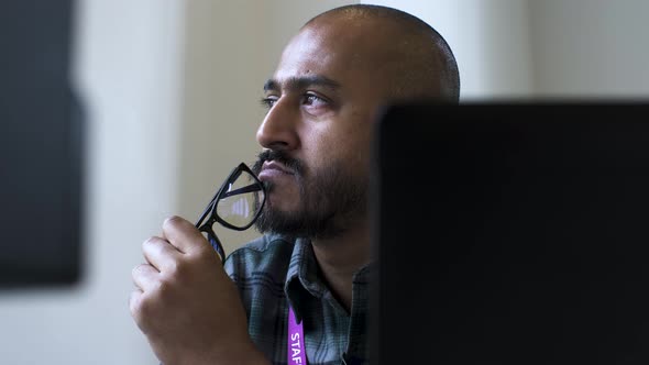 A close up shot from behind an office computer as an Asian man sits gazing into the air in deep thou