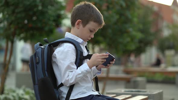 A Schoolboy Sits on a Bench in the Park and Plays on His Phone After Studying at School alt
