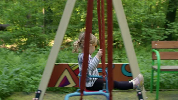 Little Blonde Girl with Ponytail in Striped Tshirt Swings on Swing