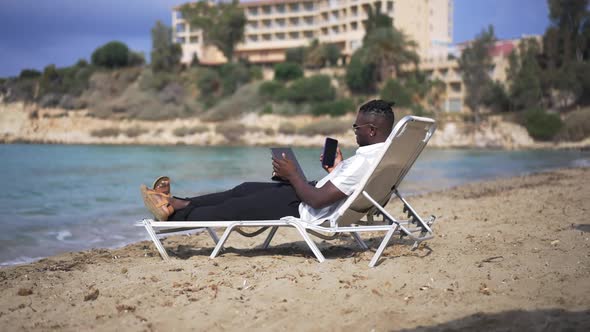 Busy African American Man Surfing Internet on Tablet Texting on Smartphone Lying on Sunbed on Sandy