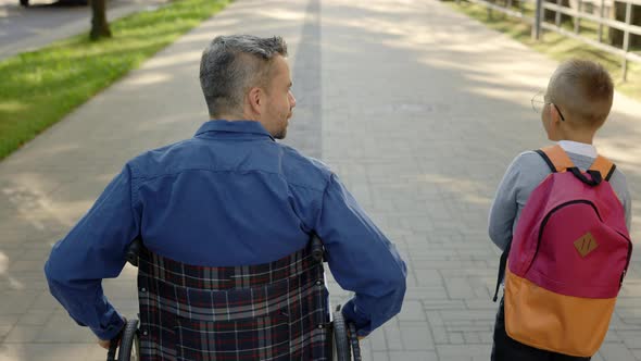 Back View of the Disabled Father in a Wheelchair and His Son Telling Him Stories From the School Son