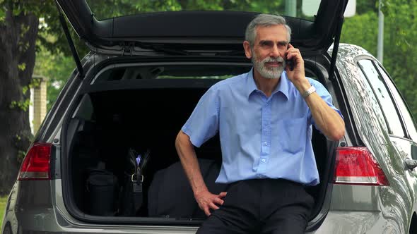 Senior Man Sits in the Trunk and Calls with the Smartphone - Trees in the Background