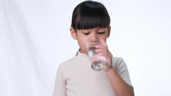 Cute little asian girl drinking water from a glass and showing thumbs up on white background in stud