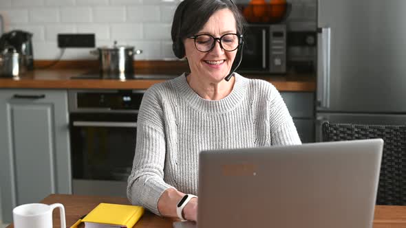 Elderly Businesswoman in Headset Speaking By Conference Call Looking at Compute