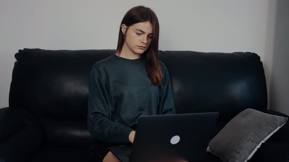 The Redhaired Focused Young Woman Opens the Laptop and Starts Typing Sitting on a Black Sofa Working