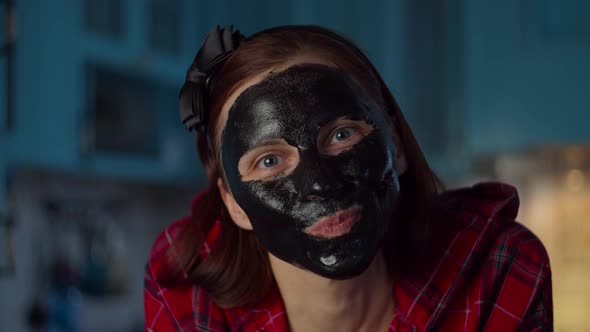 Funny 30s woman with black cosmetic mask on her face making grimace at home looking at camera