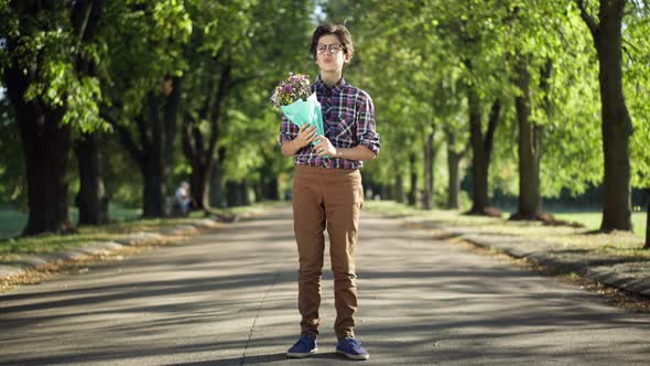 Wide Shot Anxious Teenage Boy with Bouquet of Flowers Standing in Sunny Park Looking Away