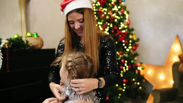 Young Happy Mother Dressed in Santa Claus Hat Helping Her Daughter to Decorate Herself for Christmas