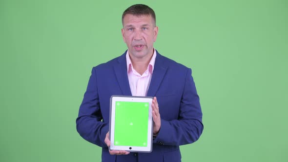 Mature Macho Businessman in Suit Talking While Showing Digital Tablet