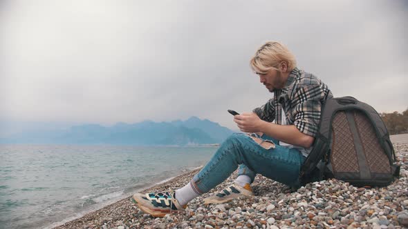 A Young Man in a Plaid Shirt Sits on the Shore and Talks on His Phone