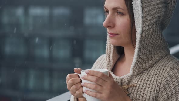 Caucasian Woman Stays on Balcony During Snowfall with Cup of Hot Coffee or Tea