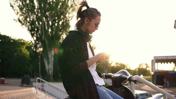 Young Girl in Black Hoodie Sitting on a Motorbike and Typing on Her Mobile