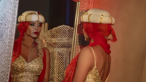 a Blonde in a Turban and a Shiny Dress Dances with a Red Scarf in Front of a Mirror