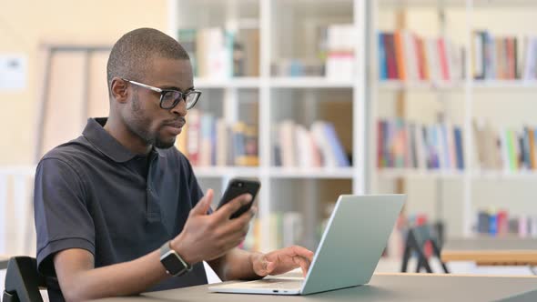 Focused African Man Working on Smartphone and Laptop in Library