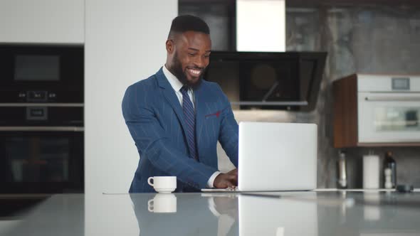 Cheerful Handsome African Businessman Wearing Suit Working with Laptop at Modern Kitchen