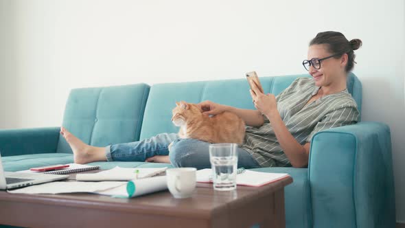 A Young Woman Lying on a Couch Holding a Smartphone and Petting a Red Cat