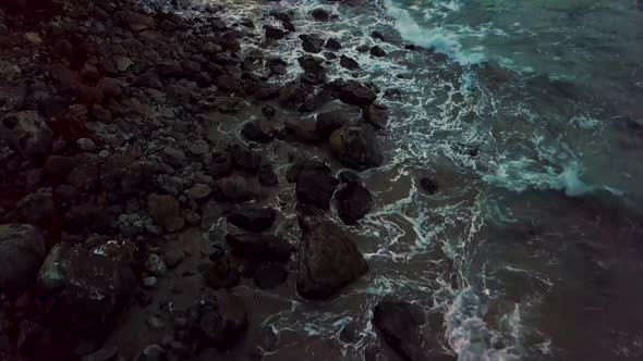 Rocky coast overhead slow reveal to sunset ocean and rolling waves crashing into huge rocks at Sand