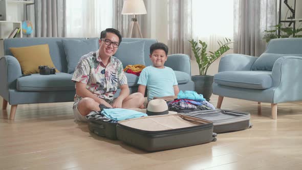 Asian Young Dad And Son Packing Clothes In Suitcase And Warmly Smile To Camera At Home
