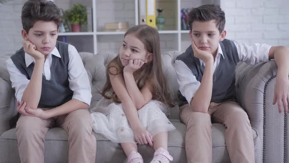 Caucasian Twin Brothers Sitting on Couch on Both Sides of Cheerful Cute Girl. Portrait of Bored