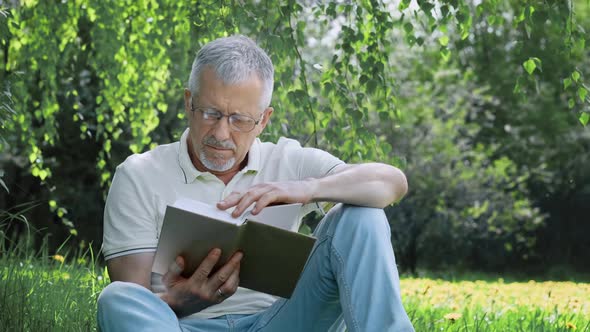 An Elderly Whitehaired Man with a Beard Sitting Alone on the Grass in a Nature Park with a Book