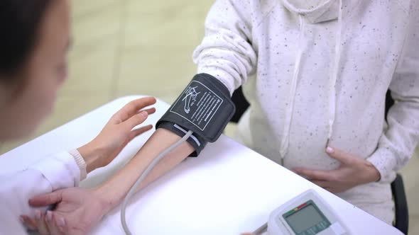 Blurred Asian Doctor Putting on Blood Pressure Gauge on Arm of Unrecognizable Pregnant Caucasian