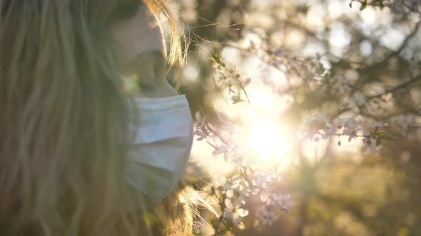 Girl in a Medical Mask Sniffs a Flowering Tree at Sunset Close-up