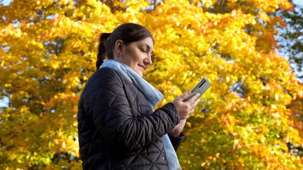 Young Woman Writes a Message on the Phone Against the Background of Yellow Trees