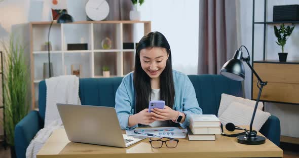 Modern Asian Girl Sitting at Her Workplace at Home and Reading Good News on Smartphone