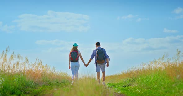 Two Tourists are Walking with Backpacks and Holding Hands Clear Blue Sky Above Their Heads