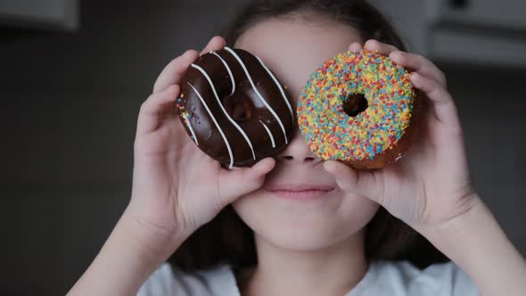 An Attractive Cute Little Girl Holding Bright Donuts with Icing in Her Hands, Smiling, Looking at